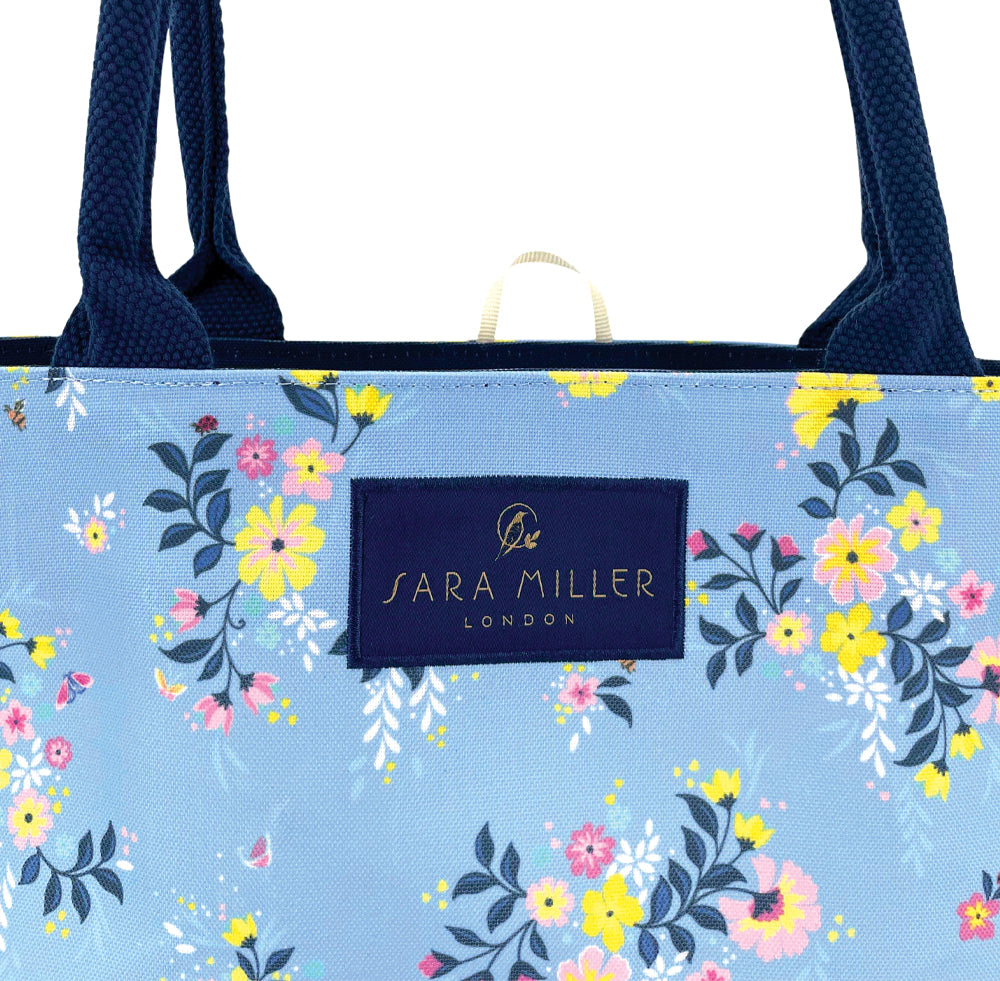 Floral-patterned tote bag with navy handles, blue bag. Close up of Sara Miller London logo.