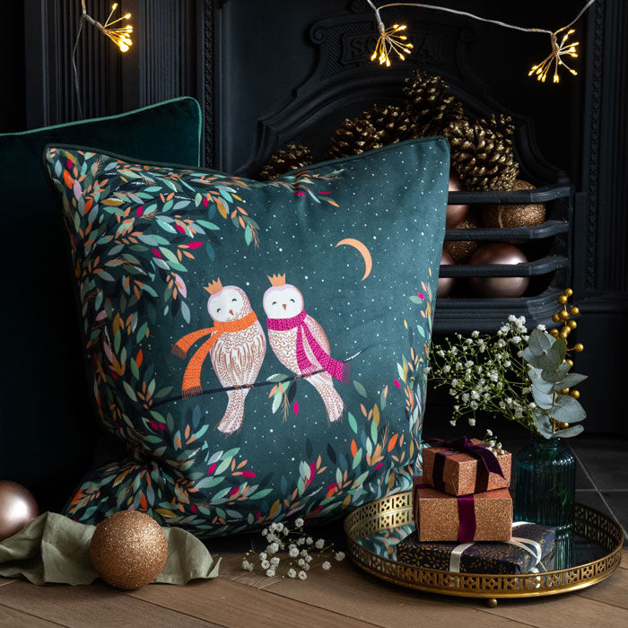A luxurious velvet festive cushion with a design of two owls perched on a branch with colourful leaves, set against a dark green background.