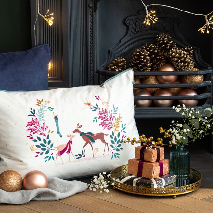 A rectangular cream velvet cushion with a festive design featuring colourful deer and foliage, and a green piping detail on the edges.
