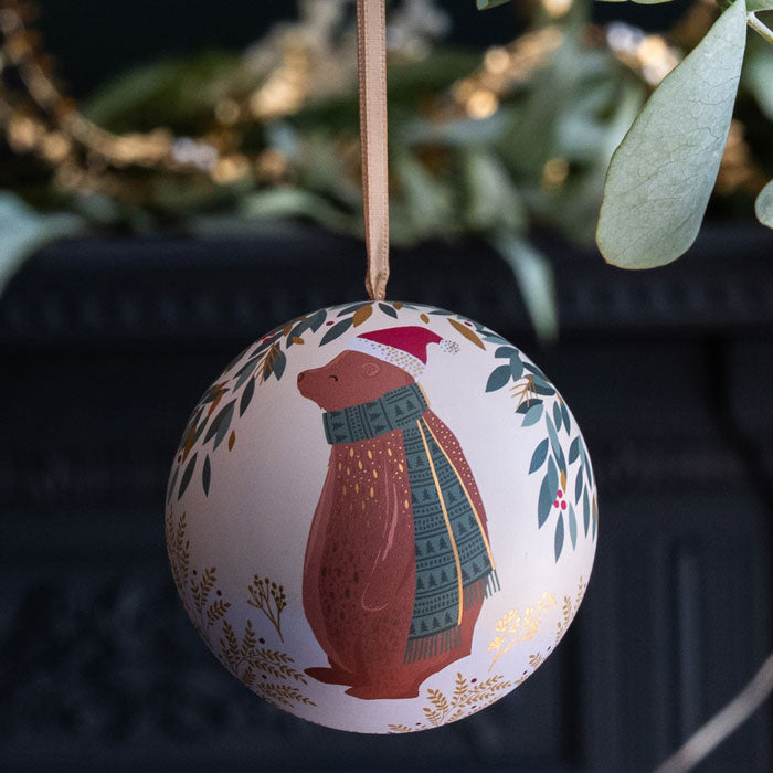 A round Christmas ornament featuring a brown bear wearing a red Santa hat and green knit scarf, hanging from a ribbon.