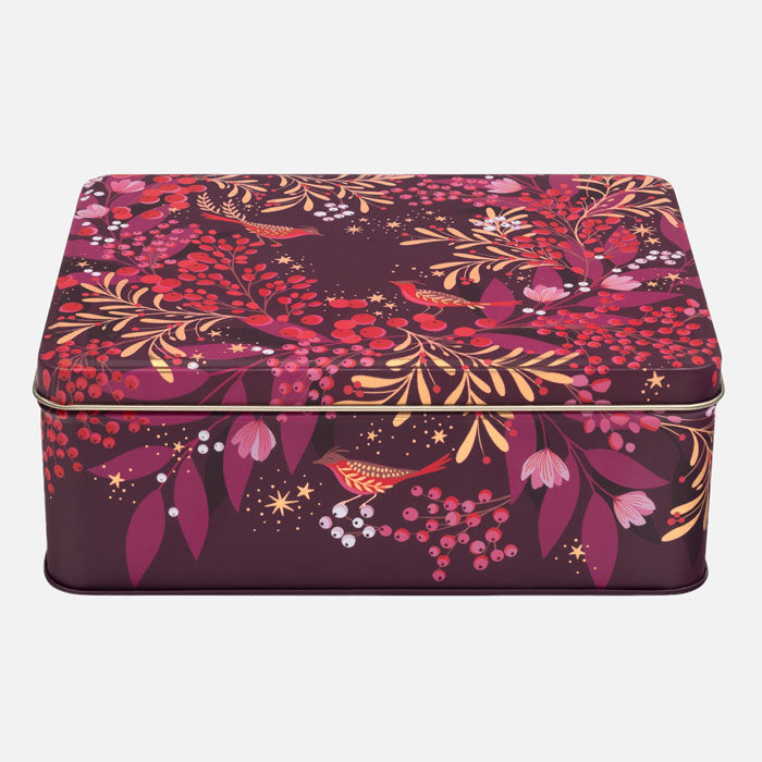 A rectangular metal tin with a lid, decorated with a festive foliage pattern featuring red berries, pink and purple leaves, gold branches, and small birds on a dark plum background.