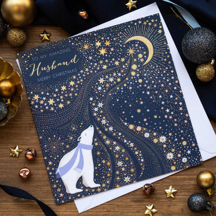 A square Christmas greeting card for Husband with a navy blue background, gold stars and crescent moon, and a white polar bear wearing a blue scarf, styled on a wooden surface with holiday ornaments and an envelope.