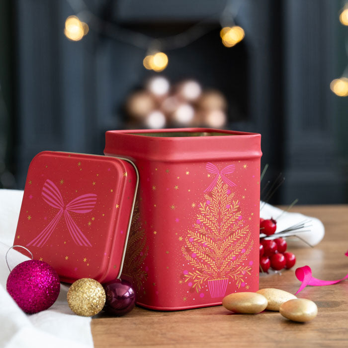 A red Christmas-themed metal tin with a gold tree design and a matching lid leaning beside it on a wooden surface, surrounded by festive ornaments and candy in a softly blurred holiday setting.