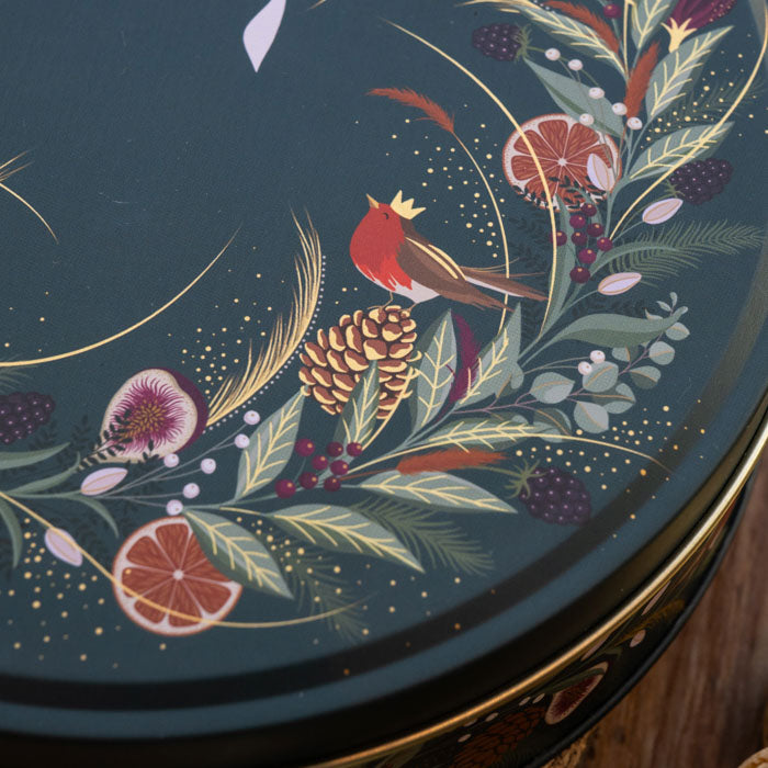 Close-up of a round decorative tin lid featuring a dark green background with hand-painted botanical and bird motifs, including a red robin, pinecone, citrus slices, berries, and gold leaf accents.