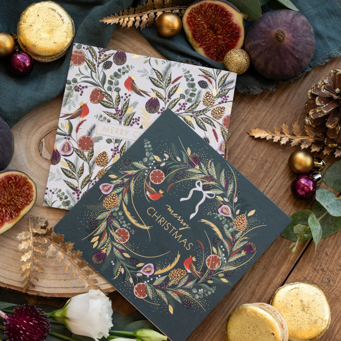Two Christmas cards styled on a wooden surface, both with festive fruits, foliage and robins. part of a mixed wallet set of cards. One green card and one cream card, with reds and greens throughout.