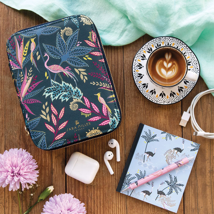 Floral patterned case, coffee cup, notebook, and earbuds on a wooden surface with a light blue cloth.