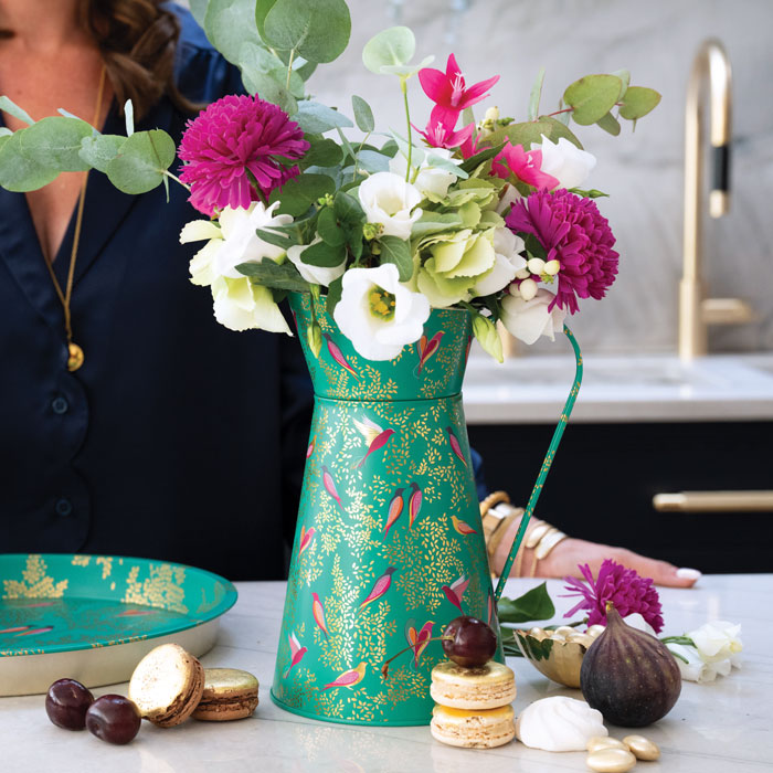 A decorative green and gold patterned tin vase filled with a colourful bouquet of white, pink, and green flowers, styled on a kitchen counter with macarons, figs, and a woman in a navy shirt partially visible in the background.