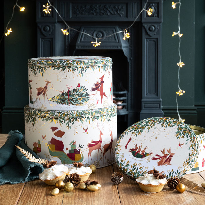 Decorative holiday tins with festive design on a wooden table in front of a fireplace.