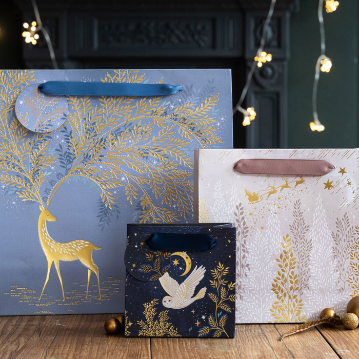 Decorative gift bags with gold tree and deer design, white owl, and gold leaves on a wooden surface.