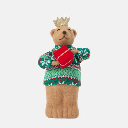 Teddy bear wearing a green sweater with red patterns and a gold crown, holding a red book on a white background.
