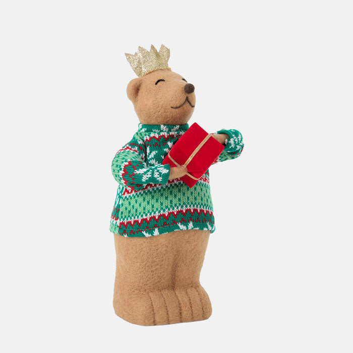 Teddy bear wearing a green sweater and gold crown, holding a red gift box on a white background