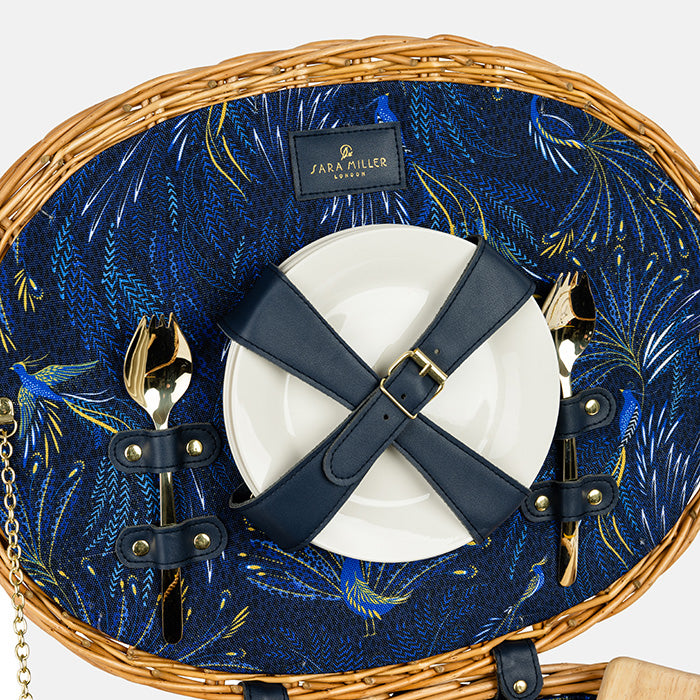 Wicker picnic basket with blue interior, white plate, black belt, and gold cutlery.