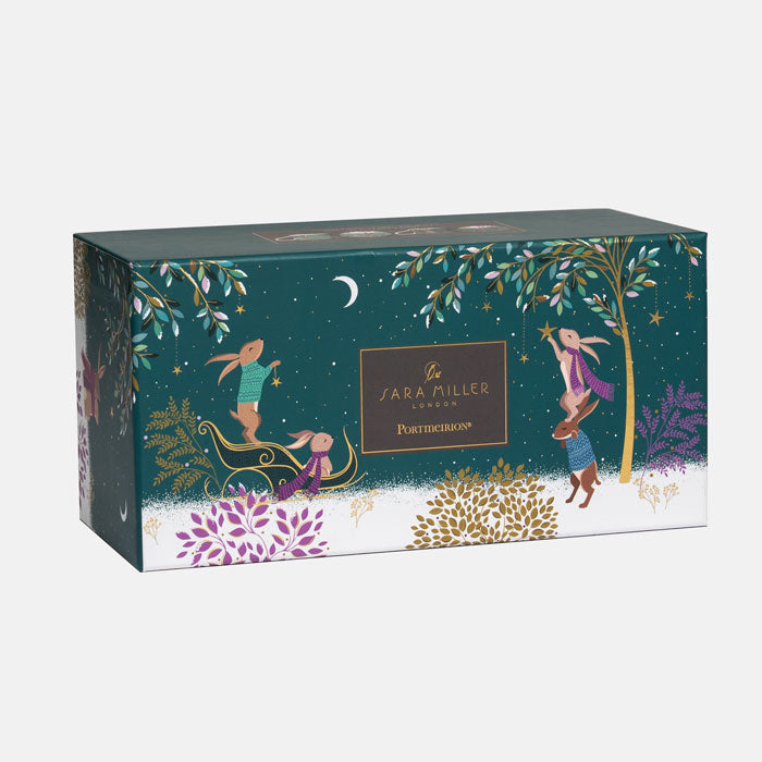 A rectangular Sara Miller London box with a whimsical winter forest illustration, featuring rabbits, a tree, stars, and purple floral accents on a dark green background.