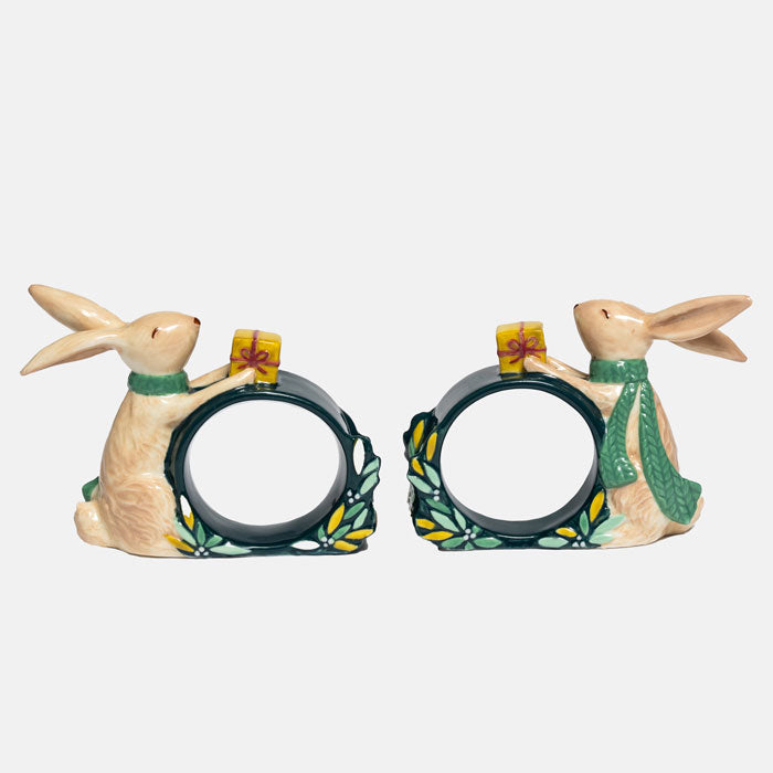 A pair of ceramic rabbit-shaped green napkin rings. The rabbits are cream-coloured with green scarves, holding yellow gift boxes, with green and yellow leafy accents at the base.