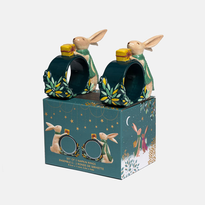 Set of two green ceramic napkin rings featuring woodland rabbits dressed in grene scarves holding yellow gifts, placed on top of their matching gift box.
