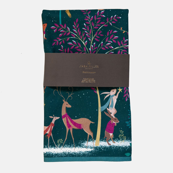 A dark green table runner with a festive Christmas woodland design, wrapped in a grey paper band. The design features deer and rabbits around a tree with pink leaves.