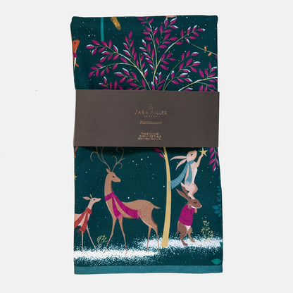 A dark green table runner with a festive Christmas woodland design, wrapped in a grey paper band. The design features deer and rabbits around a tree with pink leaves.