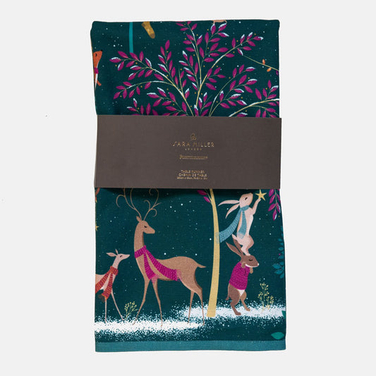 A dark green table runner with a festive Christmas woodland design, wrapped in a grey paper band. The design features deer and rabbits around a tree with pink leaves.