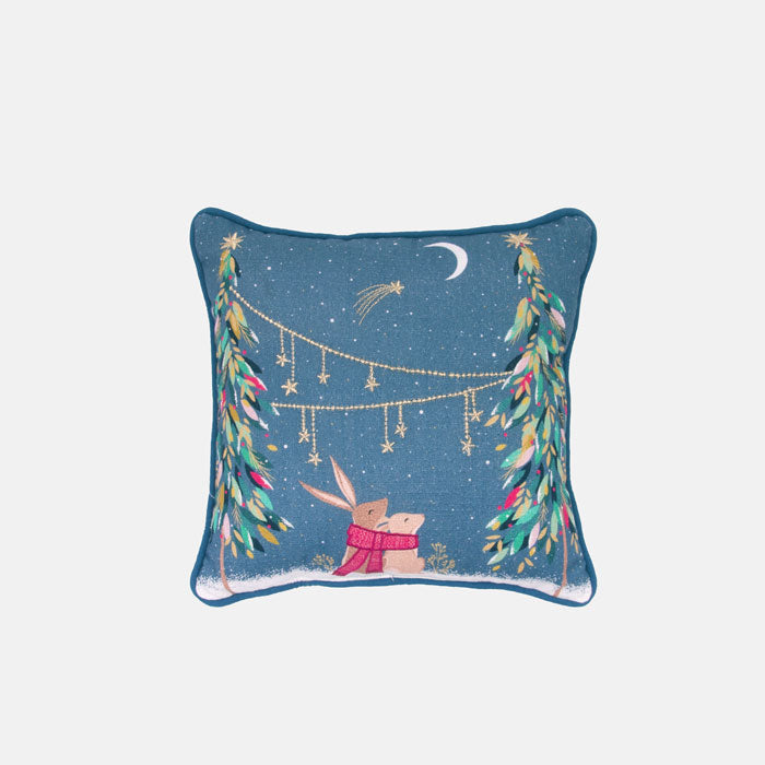 Small decorative festive cushion with rabbits sitting in moonlit scene, festive trees and starry sky, blue background.