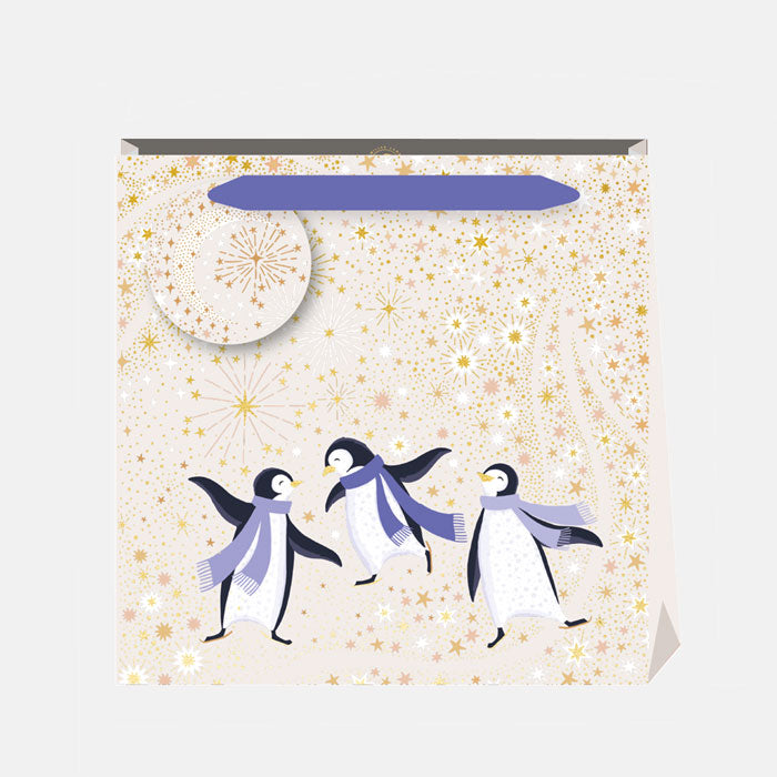A Christmas gift bag featuring penguins dancing against a starry cream and gold background, matching gift tag and purple ribbon handles.