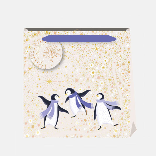A Christmas gift bag featuring penguins dancing against a starry cream and gold background, matching gift tag and purple ribbon handles.