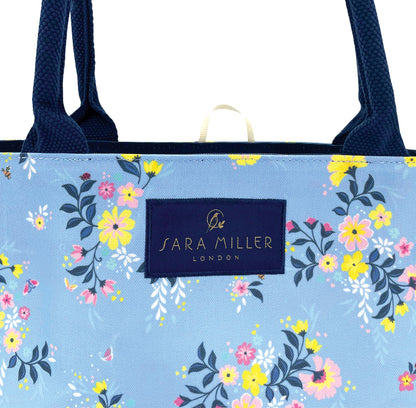 Floral-patterned tote bag with navy handles, blue bag. Close up of Sara Miller London logo.