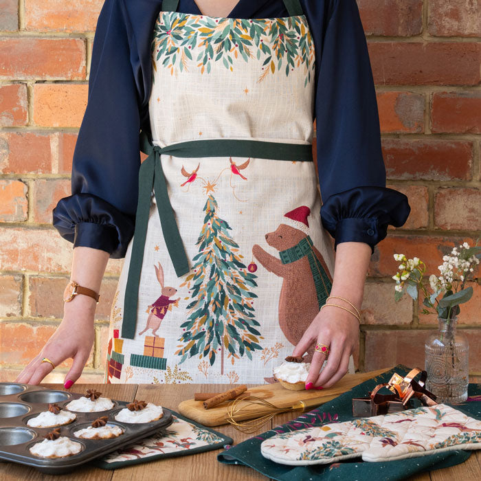 Person wearing a festive apron with a bear and tree design, standing in front of a brick wall.