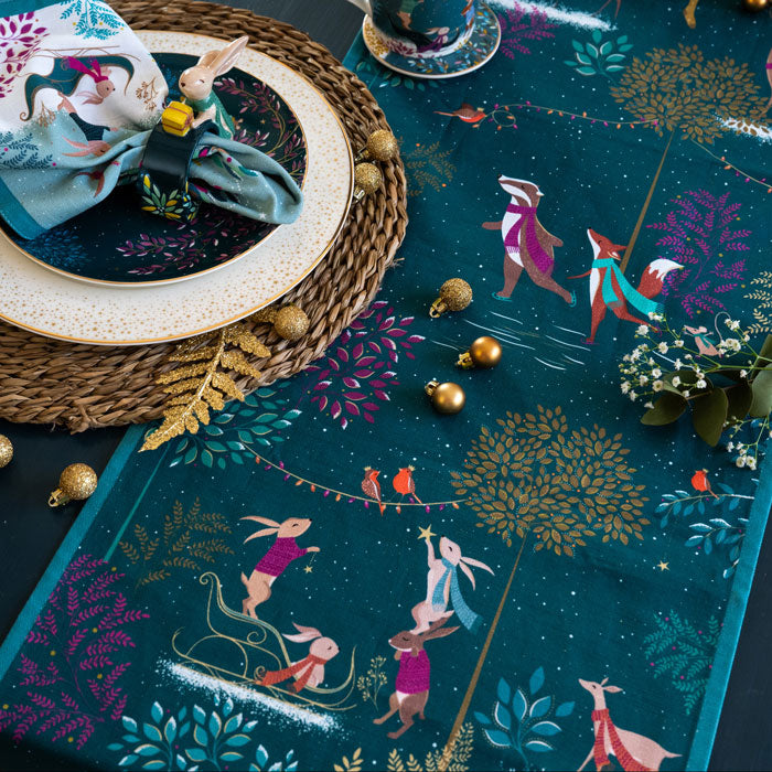 A festive dark green table runner with a whimsical woodland animal print, styled on a dark table with plates, gold ornaments, and a woven placemat.