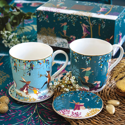 A set of two fine china mugs with matching coasters and a lid, decorated with whimsical woodland animal illustrations on green backgrounds with gold embellishment. A matching decorative gift box is visible in the background, styled on a woven mat with small flowers and gold beads.