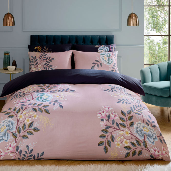 Pink floral bedding set on a bed in a bedroom with a teal chair and window in the background.