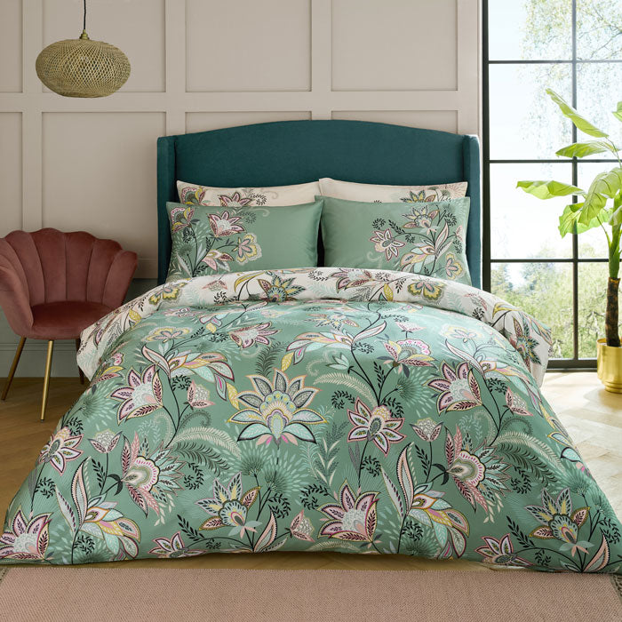 Floral bedding set in greens, creams and pinks on a bed in a bedroom with a window and plant in the background.