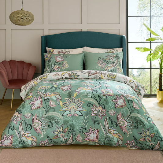 Floral bedding set in greens, creams and pinks on a bed in a bedroom with a window and plant in the background.