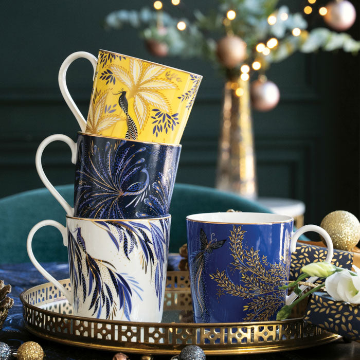 Four mugs in white, yellow, cobalt and indigo with gold embellishment all stacked on a gold tray with Christmas accents around them.