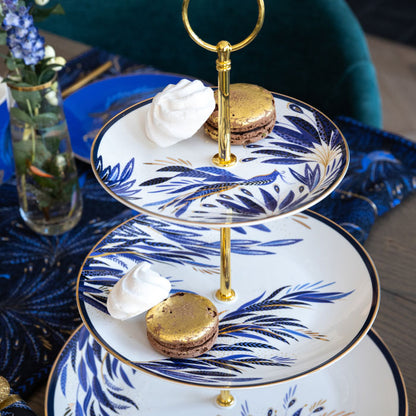 A white cake stand with indigo peacock tail feathers, gold embellishment, set on a festive table.