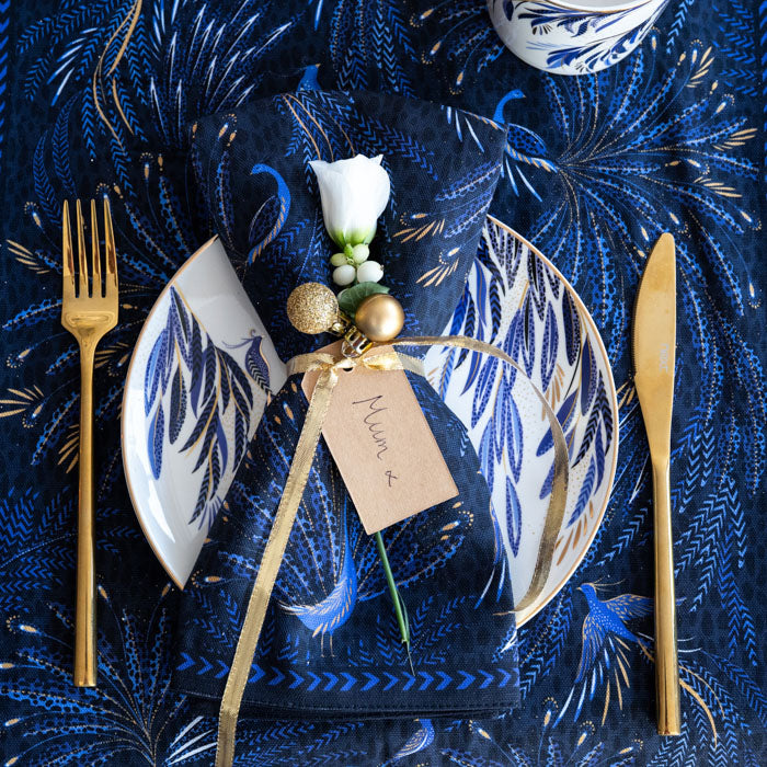 An indigo fabric napkin with peacock design set on top of a matching white plate, on a matching table runner in a festive setting.