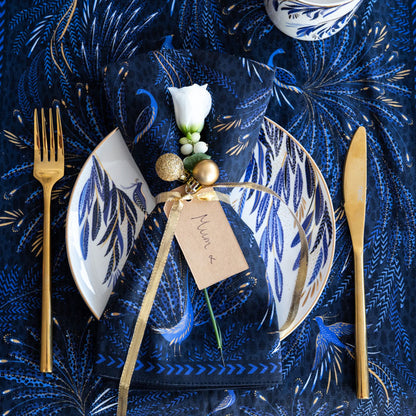 An indigo fabric napkin with peacock design set on top of a matching white plate, on a matching table runner in a festive setting.