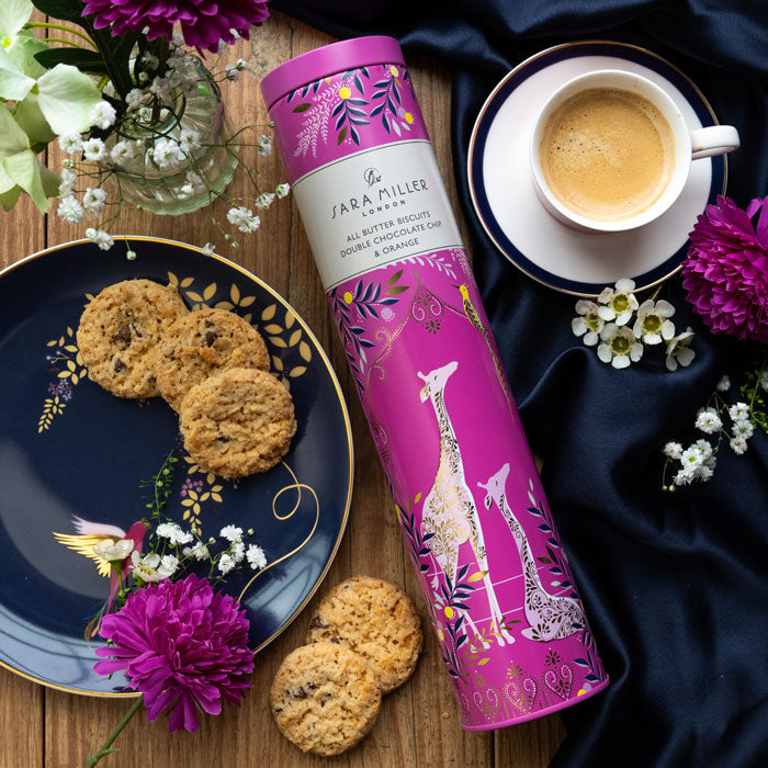 Pink tea canister with giraffe design on a table with cookies, coffee, and flowers.