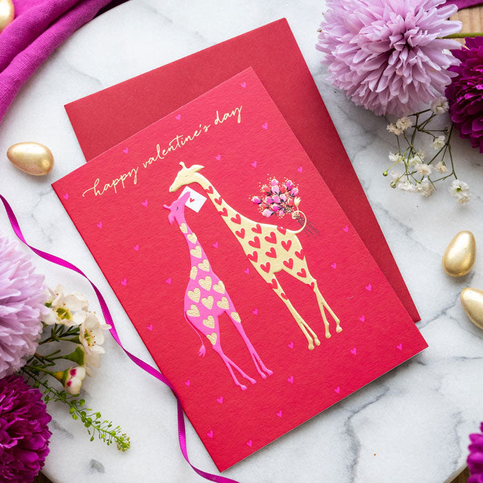 Valentine's Day card with two giraffes holding flowers on a red background, surrounded by flowers and ribbons.