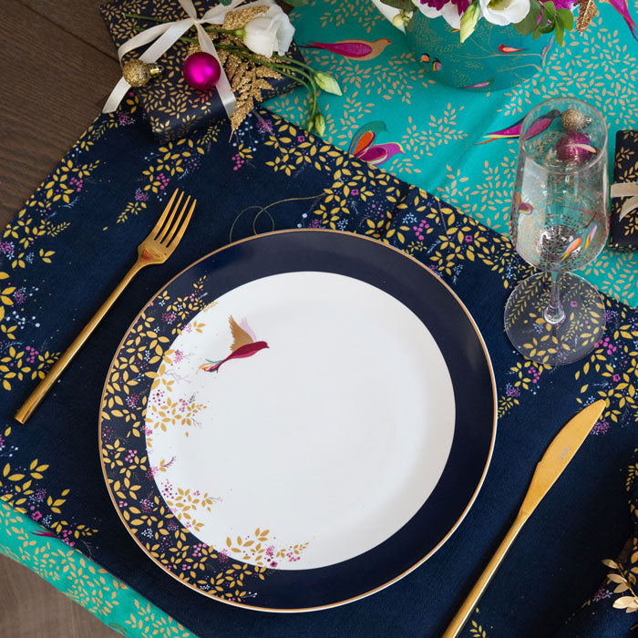 A navy blue fabric placemat with a hummingbird design and foliage, set on a table with matching plate.