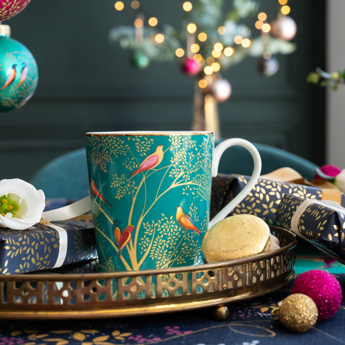 A green mug with birds and branches design, set on a gold tray surrounded by Christmas accents.