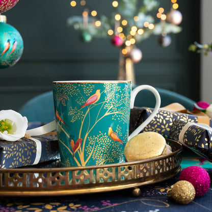 A green mug with birds and branches design, set on a gold tray surrounded by Christmas accents.