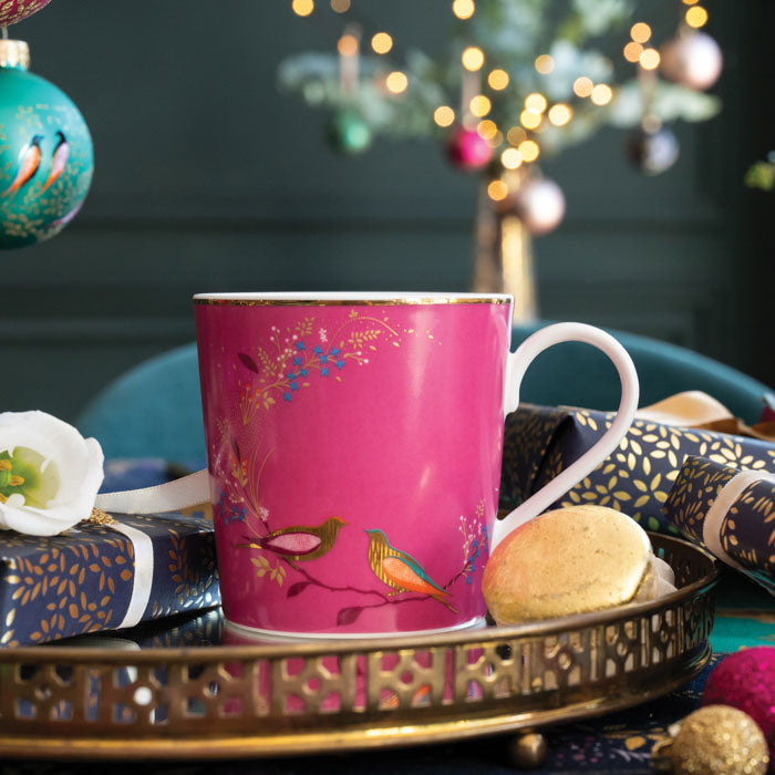 A pink china mug with birds resting on a branch, gold embellishment, surrounded by Christmas accents.