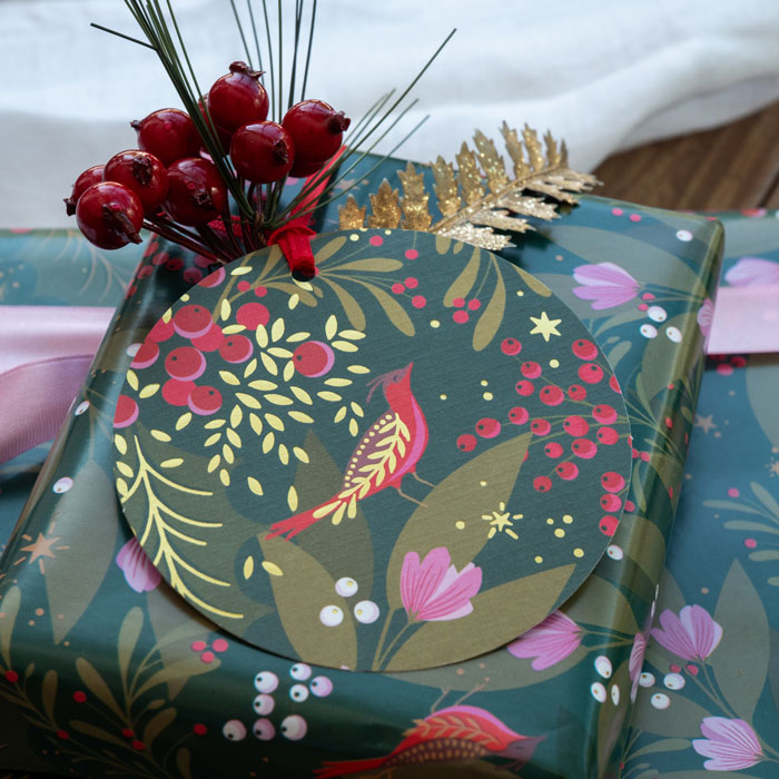 A green gift tag with a festive pattern with berries, robin and foliage in pink and red colours, attached to a present with matching wrapping paper.