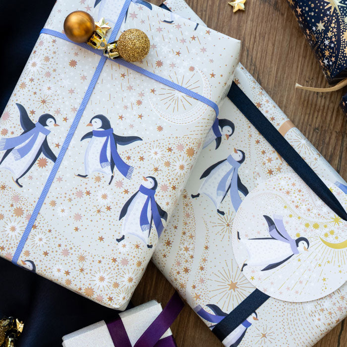 A cream gift tag with little penguin dressed in purple scarf, under a crescent moon and starry sky attached to a present with matching wrapping paper.