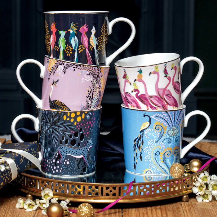 Five colourful mugs stacked on a gold tray, with Christmas baubles and wrapped presents all around.