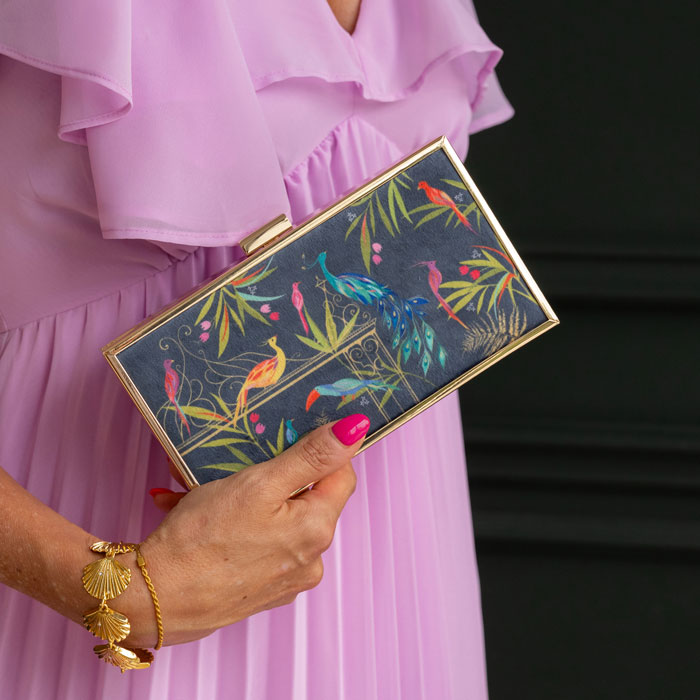 Hand holding a colourful clutch with floral and bird design, wearing a pink dress.