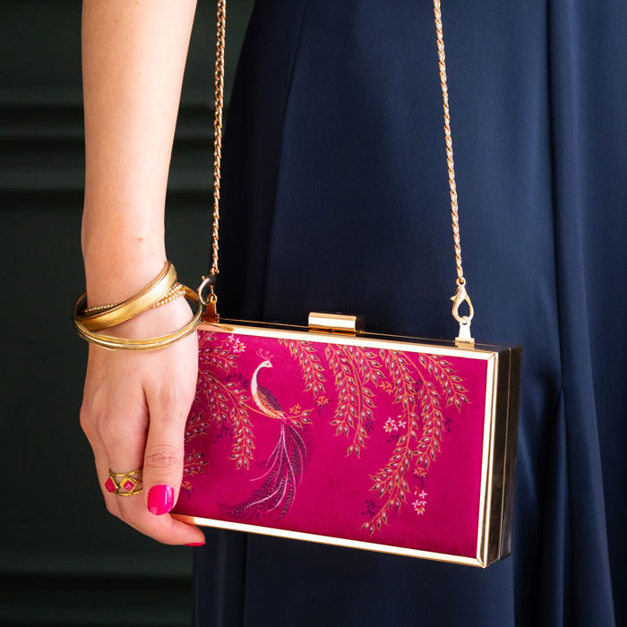 Pink clutch with gold accents held by a hand wearing gold bangles against a dark background
