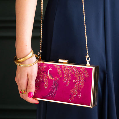 Pink clutch with gold accents held by a hand wearing gold bangles against a dark background