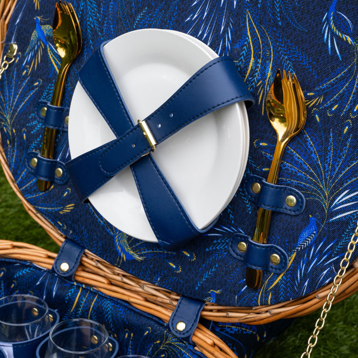 Picnic basket with blue straps, white plate, gold cutlery, and blue floral fabric background