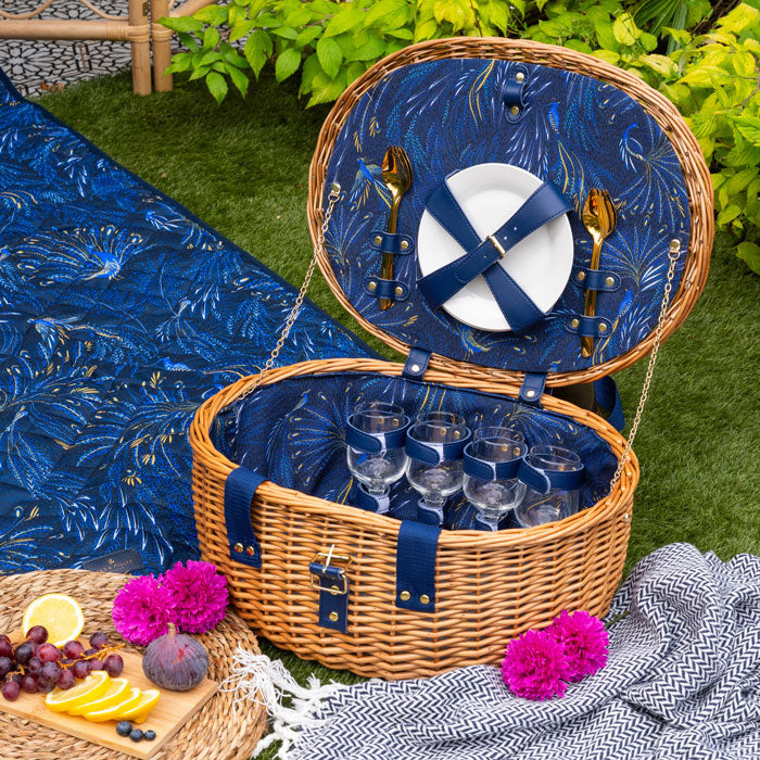 Open picnic basket with blue interior, cutlery, and glasses on a grassy area with a blue blanket and food items.